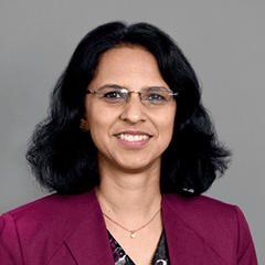 Megha Budruk headshot - medium-skinned woman, glasses, smiling, black hair, burgundy blazer, necklace