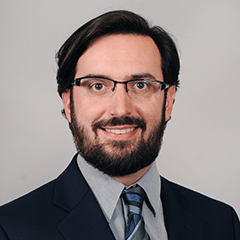 David Androff headshot - fair-skinned man, smiling, black hair and beard, glasses, gray collared shirt, black blazer, blue striped tie